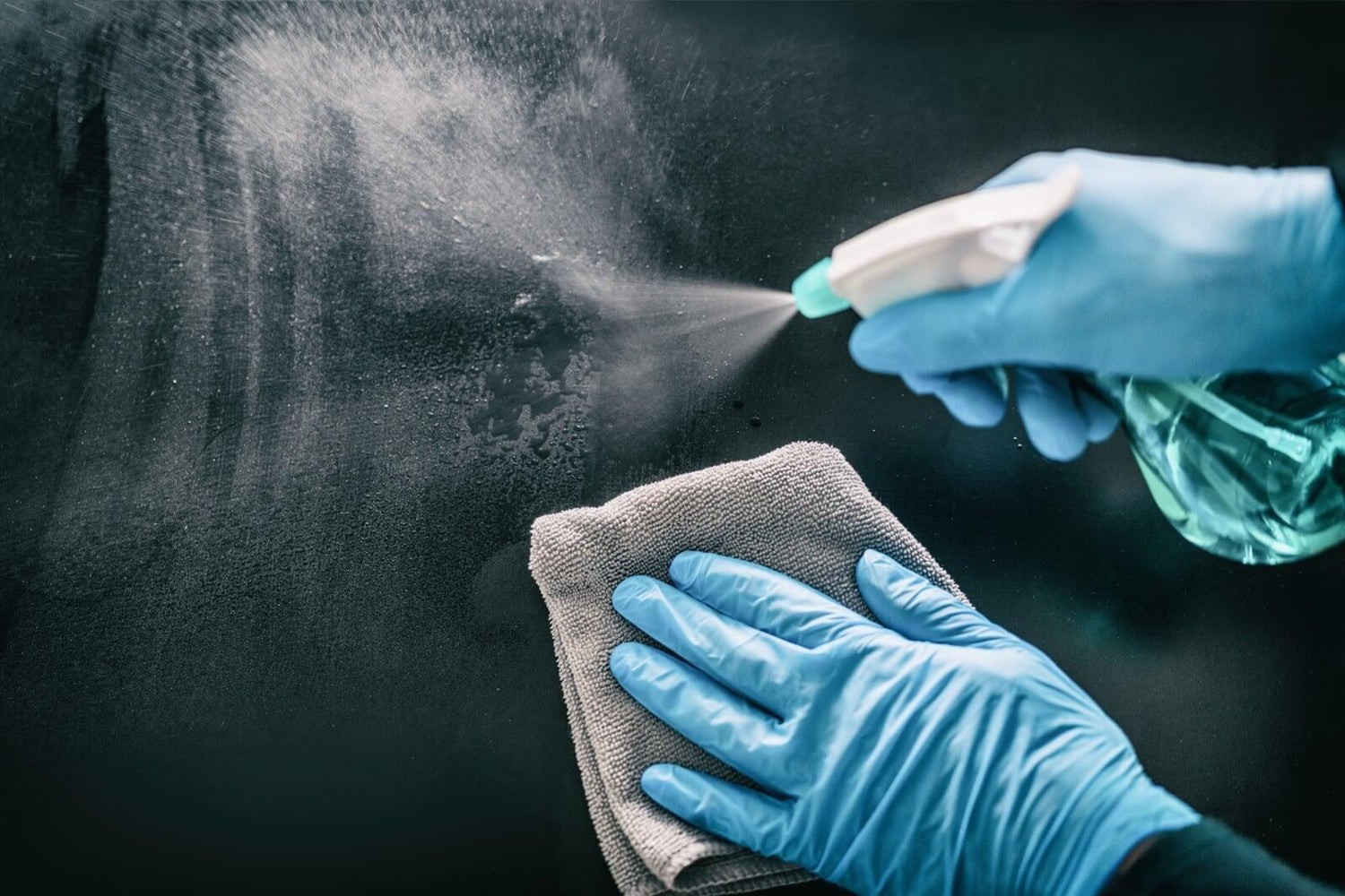 A person wearing blue gloves sprays and wipes down a glass surface with a grey cloth and glass cleaner.